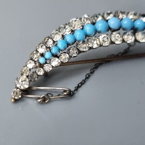 May include: A silver and gold brooch with a curved design. The brooch is decorated with turquoise and clear rhinestones. The brooch has a safety pin closure and a chain.