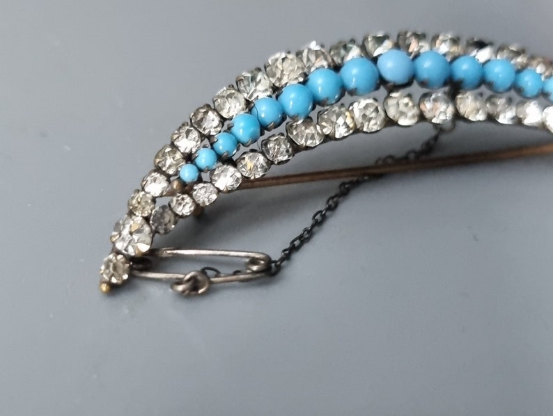 May include: A silver and gold brooch with a curved design. The brooch is decorated with turquoise and clear rhinestones. The brooch has a safety pin closure and a chain.