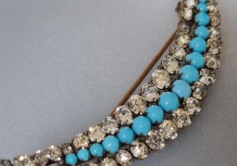 May include: A silver and turquoise brooch with a crescent shape. The brooch is made of small, faceted stones and features a row of turquoise stones.