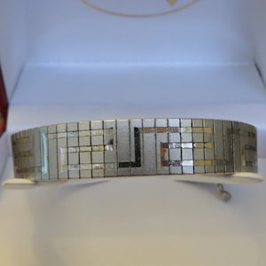 May include: A silver-toned bracelet with a geometric pattern, presented in a red and white box. The bracelet features a repeating square and rectangular design. The jewelry is displayed on a white stand.