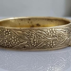 Pode incluir: Um bracelete dourado com um design floral intrincado. O bracelete é articulado e tem uma corrente presa ao fecho.