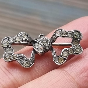 以下が含まれることがあります： リボンの形をしたシルバー調の金属製ブローチ。リボンのループと中央の結び目全体に、小さな透明な丸いラインストーンが埋め込まれており、裏側には水平なピンのメカニズムが見えます。
