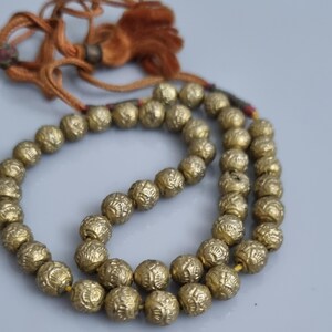 May include: A gold beaded necklace with a tassel. The beads are round and have a textured surface. The necklace is strung on a brown cord.