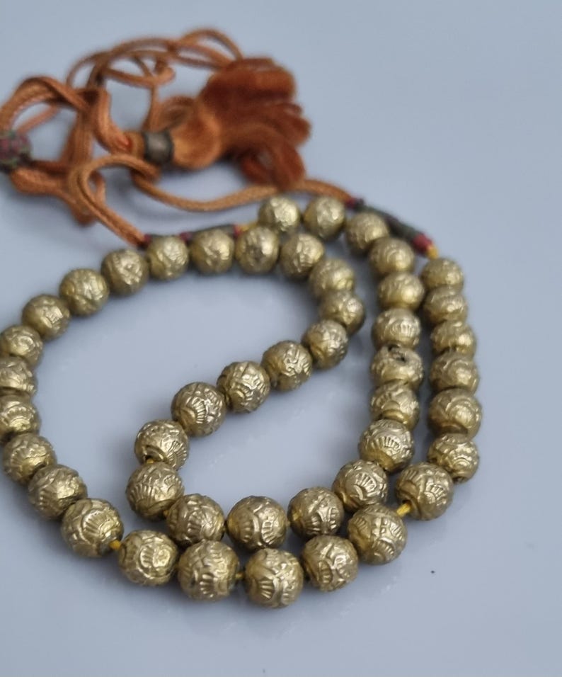 May include: A gold beaded necklace with a tassel. The beads are round and have a textured surface. The necklace is strung on a brown cord.