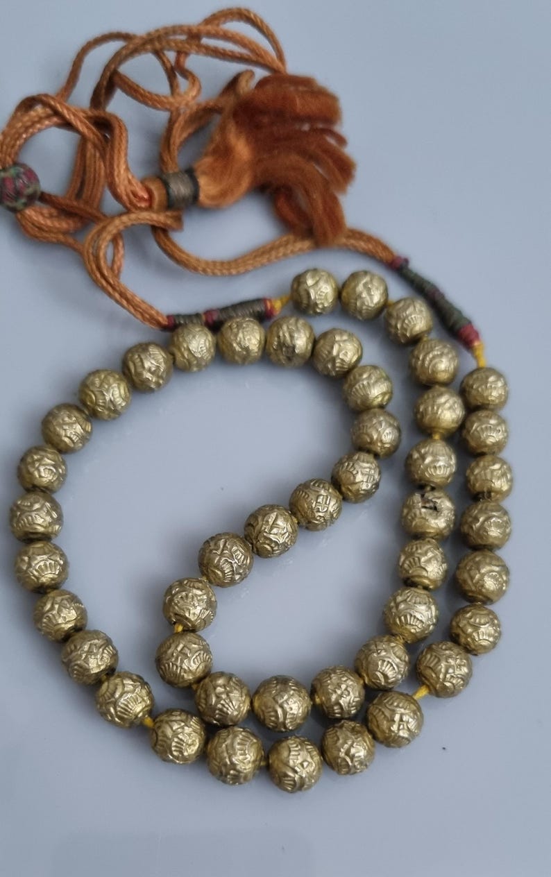 May include: A gold beaded necklace with a tassel. The beads are round and have a textured surface. The tassel is made of brown cord.
