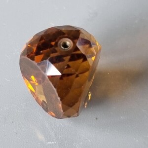 May include: A faceted, amber-colored glass bead with a small hole in the top. The bead has a unique, irregular shape.
