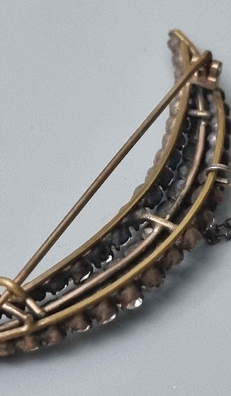 May include: A crescent moon shaped brooch with a gold tone metal setting and a row of clear rhinestones around the edge.