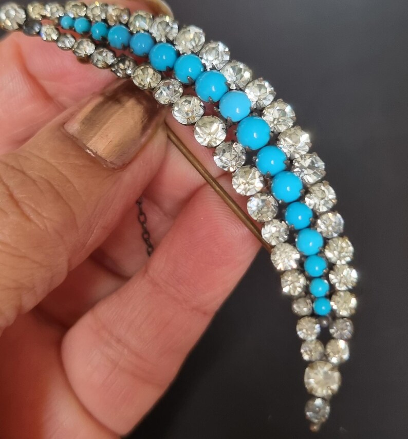 May include: A vintage brooch with a crescent shape design featuring a row of turquoise blue stones surrounded by clear rhinestones.