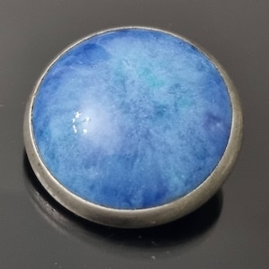 Pode incluir: Um cabochão redondo azul e branco engastado em uma moldura de prata. O cabochão tem uma aparência marmorizada e nublada.