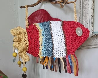 Può includere: Un pesce appeso al muro in crochet con un corpo a strisce rosso, bianco, giallo, blu e marrone. Il pesce ha una coda in crochet gialla con perline e una pinna in pelle marrone. Il pesce è appeso a un ramo.