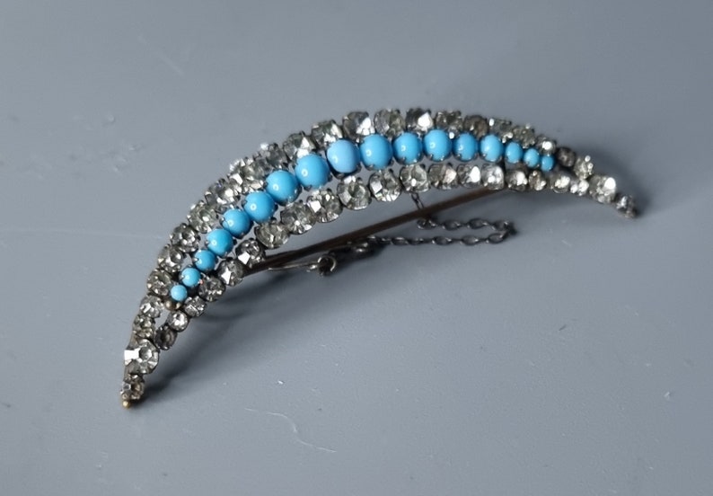 May include: A crescent-shaped brooch with a row of turquoise blue stones set in silver, surrounded by clear rhinestones.