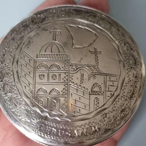 May include: A silver circular box with an engraved design of a church building with two crosses on top. The inscription "JERUSALEM" is engraved on the bottom of the box.