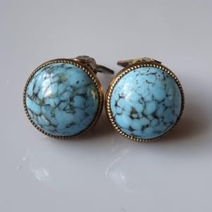 May include: A pair of round cufflinks with a light blue, marbled stone set in a gold-colored metal frame. The cufflinks have a textured, rope-like border and a silver-colored clasp.