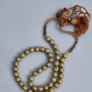 May include: A gold beaded necklace with a tassel. The beads are round and have a textured surface. The necklace is strung on a brown cord.