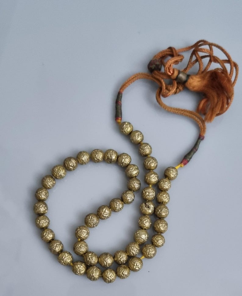 May include: A gold beaded necklace with a tassel. The beads are round and have a textured surface. The necklace is strung on a brown cord.