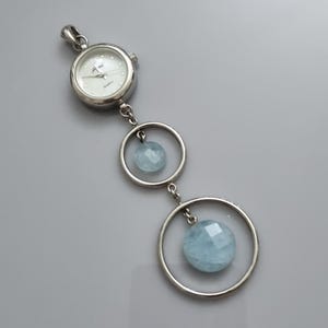 May include: A silver watch with a white face and a pendant with two blue aquamarine stones hanging from it.