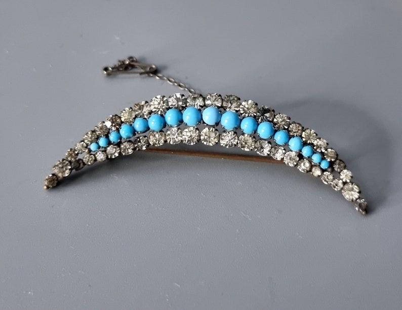 May include: A crescent-shaped brooch with a row of turquoise blue stones surrounded by clear rhinestones.