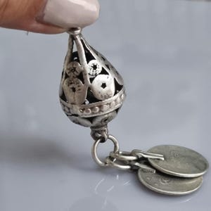 May include: Silver pendant in a teardrop shape, featuring an openwork design and small coin charms. The pendant is suspended from a ring and has a detailed, textured surface. The coin charms hang below, adding movement and visual interest.