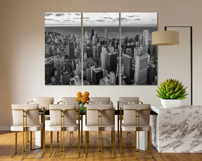 May include: A black and white photo of a city skyline with tall buildings. The photo is split into three panels and is hanging on a wall above a dining table with chairs.