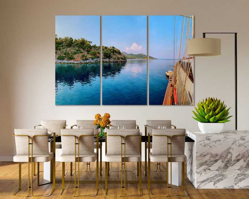 May include: A three-panel canvas print depicting a view of a blue body of water with a green island in the distance. The right panel shows a wooden boat with white sails.