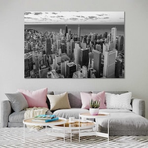 May include: A black and white aerial photograph of a city skyline. The buildings are tall and clustered together, with a few taller buildings standing out. The sky is cloudy and the water in the distance is calm.