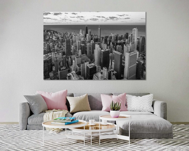 May include: A black and white aerial photograph of a city skyline. The buildings are tall and clustered together, with a few taller buildings standing out. The sky is cloudy and the water in the distance is calm.