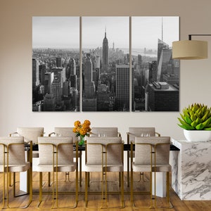 May include: Black and white photograph of the New York City skyline, featuring the Empire State Building and other skyscrapers. The photo is split into three panels and is displayed on a wall above a dining table with chairs.