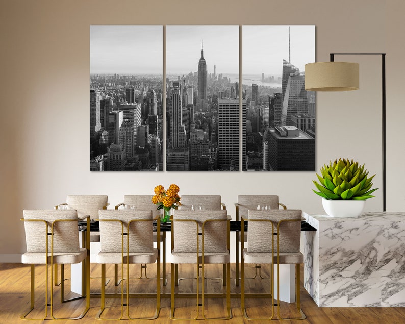 May include: Black and white photograph of the New York City skyline, featuring the Empire State Building and other skyscrapers. The photo is split into three panels and is displayed on a wall above a dining table with chairs.