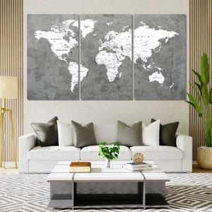 May include: A three-panel canvas print of a world map with a grey background and white lines. The map is divided into three sections, with North America, Europe, and Asia on the left panel, Africa and the Middle East on the centre panel, and South America, Australia, and Antarctica on the right panel. The map is displayed on a white wall above a white sofa with grey cushions.