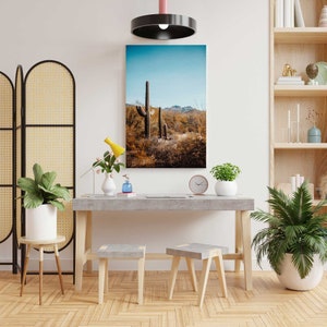 May include: A minimalist living room interior with a concrete desk, two wooden stools, and a framed print of a desert landscape with cacti. The room is decorated with plants and a screen.