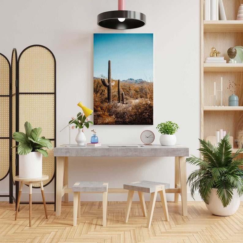 May include: A minimalist living room interior with a concrete desk, two wooden stools, and a framed print of a desert landscape with cacti. The room is decorated with plants and a screen.