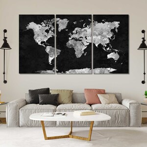 May include: A black and white world map print in a three-piece set. The map is a distressed, textured design with a white outline. The map is displayed on a white wall above a grey sofa with cushions and a coffee table.