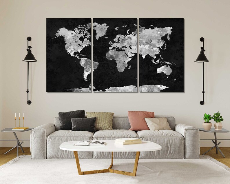 May include: A black and white world map print in a three-piece set. The map is a distressed, textured design with a white outline. The map is displayed on a white wall above a grey sofa with cushions and a coffee table.