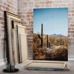 May include: A set of four framed canvases with a desert landscape featuring a tall cactus in the foreground. The canvases are stacked on top of each other and are leaning against a brick wall.