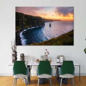 Puede incluir: Una fotograf&iacute;a panor&aacute;mica de los Acantilados de Moher en Irlanda, con un cielo de puesta de sol dram&aacute;tico y un afloramiento rocoso en el oc&eacute;ano. Los acantilados son verdes y marrones, y el agua es de un azul profundo.