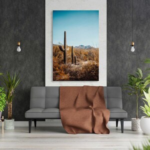 May include: A photograph of a desert landscape with three tall cacti in the foreground. The cacti are brown and green and are set against a blue sky with white clouds. The photo is framed and hanging on a wall in a living room setting.