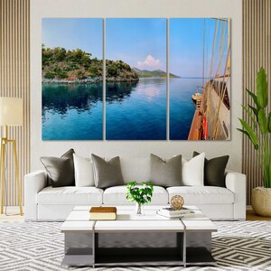 May include: A panoramic view of a sailboat on the water with a small island in the background. The water is a deep blue and the sky is a light blue. The sailboat is white with a brown sail.