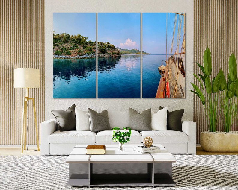 May include: A panoramic view of a sailboat on the water with a small island in the background. The water is a deep blue and the sky is a light blue. The sailboat is white with a brown sail.