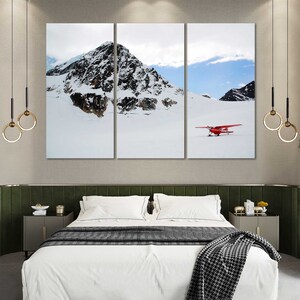 May include: A three-panel canvas print featuring a snowy mountain landscape with a red airplane flying over the snow-covered ground. The print is displayed in a modern bedroom setting with a green headboard and a white bed.