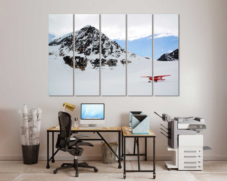 May include: A multi-panel canvas print of a snowy mountain landscape with a red airplane flying over the snow. The print is hanging on a white wall above a desk with a computer, chair, and printer.