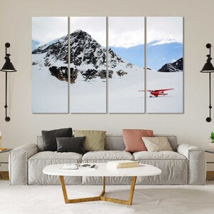 May include: A red airplane sits on a snowy mountain landscape. The plane is in the foreground, with a snow-covered mountain range in the background. The sky is blue with white clouds.
