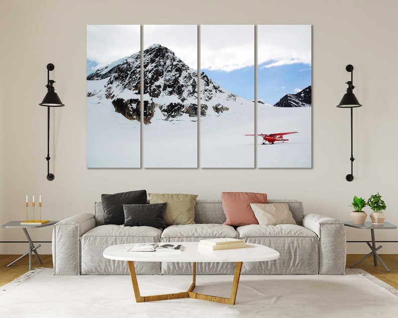 May include: A red airplane sits on a snowy mountain landscape. The plane is in the foreground, with a snow-covered mountain range in the background. The sky is blue with white clouds.