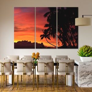May include: A tri-panel canvas print featuring a sunset over a tropical island with palm trees silhouetted against the orange and pink sky. The print is displayed above a dining table with chairs.