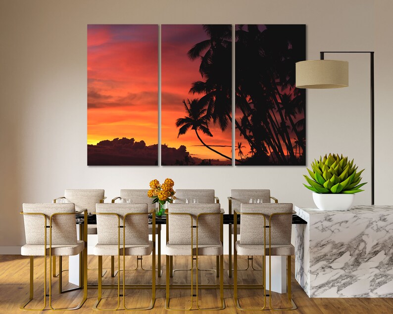 May include: A tri-panel canvas print featuring a sunset over a tropical island with palm trees silhouetted against the orange and pink sky. The print is displayed above a dining table with chairs.