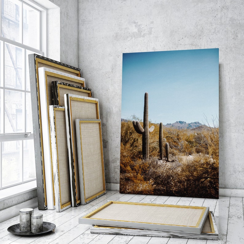 May include: A stack of four canvas frames leaning against a white wall. The frames are different sizes and are made of wood with a natural finish. The largest frame is empty, while the other three frames are filled with canvas. The frames are in a room with a large window and a concrete floor. A large print of a desert landscape with a cactus in the foreground is hanging on the wall.