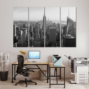 May include: A black and white photo of a city skyline, split into three panels. The photo is hanging on a white wall above a desk with a black office chair, a computer, and a printer.