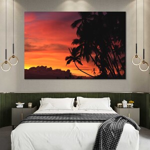 May include: A bedroom interior with a large framed print of a sunset over palm trees. The print is hanging above a bed with white linens and a black and white throw blanket. There are two hanging light fixtures with gold rings.