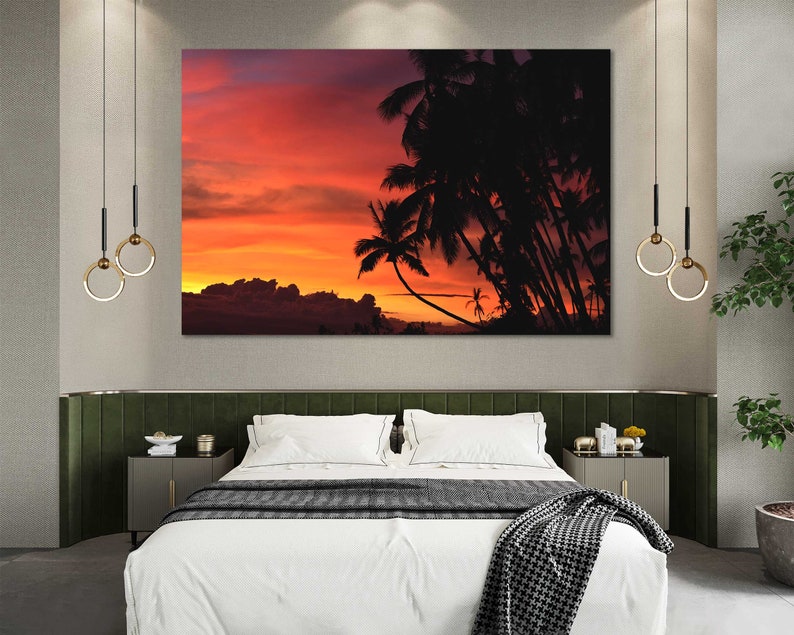 May include: A bedroom interior with a large framed print of a sunset over palm trees. The print is hanging above a bed with white linens and a black and white throw blanket. There are two hanging light fixtures with gold rings.