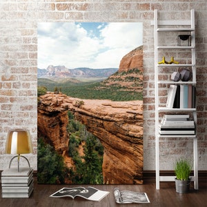 May include: A large canvas print depicting a natural rock bridge in a canyon landscape. The image shows red rock formations, green trees, and a blue sky with clouds. The print is displayed in a room with a brick wall and a white bookshelf.