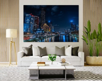 May include: A large canvas print of a nighttime cityscape, featuring illuminated skyscrapers reflected in a river. The artwork hangs above a white sofa with gray pillows, complemented by a modern coffee table and a floor lamp.